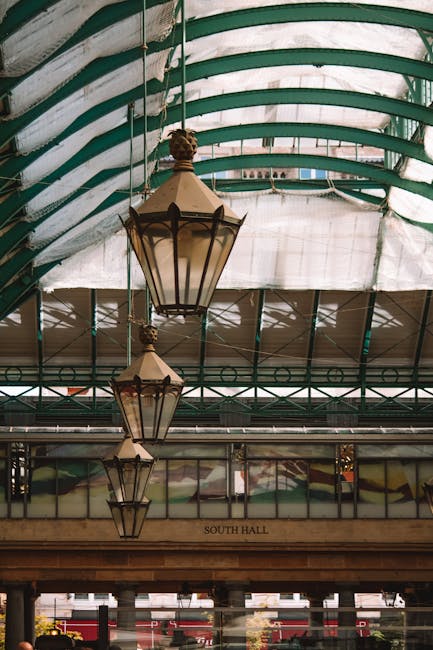 An interior view of Covent Garden's South Hall featuring a glass and metal roof with visible green framing and striped fabric panels. Hanging from the ceiling are three large, vintage-style lanterns with metal frames and frosted glass panels, arranged in a descending size. Below the lanterns, a wooden beam with the inscription 'South Hall' is visible, along with glass windows that reveal outdoor areas. The environment suggests a historic or market building characteristic of busy European market places, where furniture or boxes could be transported during home relocation or moving processes, as managed by companies like Man and Van Covent Garden.