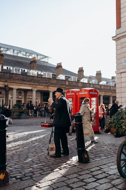A street scene in Covent Garden with a man dressed in a black suit and bowler hat holding a coffee cup, standing on a cobblestone pavement. Nearby, a woman in a beige coat walks past a red traditional British telephone booth, while other pedestrians are visible in the background. The area is busy with people and flower stalls, and the rooftops of historic buildings with chimneys and a glass-roofed structure can be seen above. The scene depicts an urban environment during daytime, illustrating typical street activity and pedestrian movement associated with home relocation or moving logistics in central London. Man and Van Covent Garden operates in this area for furniture transport and packing and moving services.