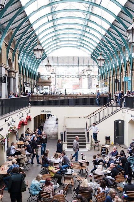 An interior view of Covent Garden's South Hall featuring a glass and metal roof with visible green framing and striped fabric panels. Hanging from the ceiling are three large, vintage-style lanterns with metal frames and frosted glass panels, arranged in a descending size. Below the lanterns, a wooden beam with the inscription 'South Hall' is visible, along with glass windows that reveal outdoor areas. The environment suggests a historic or market building characteristic of busy European market places, where furniture or boxes could be transported during home relocation or moving processes, as managed by companies like Man and Van Covent Garden.