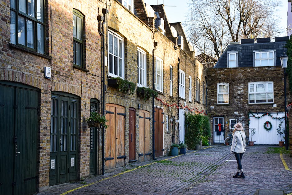 A woman dressed in a light coat and hat stands on a cobblestone alleyway in a residential area of Covent Garden, surrounded by brick houses with clapboard and brick facades, some featuring window boxe