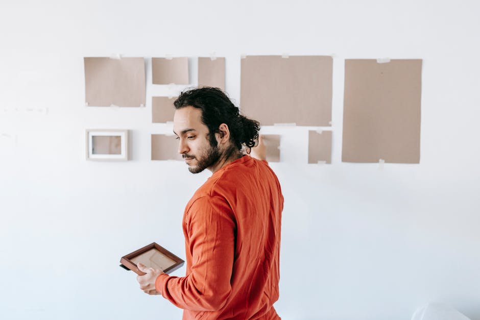 A man with dark, curly hair, dressed in an orange long-sleeved shirt, stands in front of a white wall with several blank picture frames and canvases arranged in a gallery-style display. He is holding 