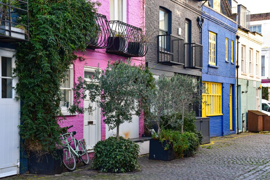 A row of residential buildings with brick and painted facades along a cobbled street, featuring small balconies with black metal railings, potted plants in various containers, and closed windows with