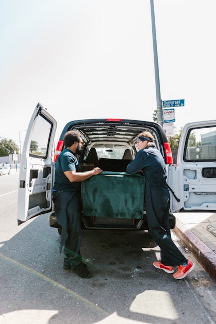 Two movers from Man and Van Covent Garden are engaged in a home relocation task, standing at the open rear of a black van parked on a street in Covent Garden. They are handling a large, green upholstered piece of furniture, which is partially wrapped in protective fabric or felt, and are carefully lifting it into the vehicle's cargo space. Surrounding the van, there are typical urban elements such as a lamp post with a street sign, pavement, and nearby parked cars, indicating a tight loading bay environment. Inside the van, visible cardboard and plastic wrapping materials are used for packing and safeguarding belongings during furniture transport, while a trolley or dolly might be implied for aiding the heavy lifting, although not directly visible. The scene captures the precise moment of loading furniture securely in preparation for a house move or relocation, emphasizing the importance of careful handling within the logistical process of packing and moving in urban settings, consistent with professional removals services.